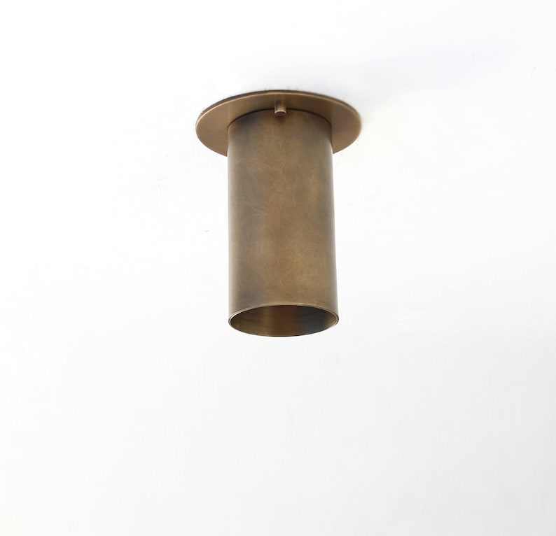May include: A cylindrical ceiling light fixture with a brushed brass finish. The fixture features a flat circular mounting plate at the top that connects to a vertical tube, creating a simple, functional design for overhead lighting installations.