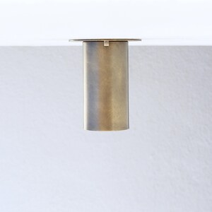 May include: A cylindrical furniture leg with a brushed brass finish. The leg features a flat circular mounting plate at the top, designed for attachment to a flat surface. The metal surface has a muted, matte texture with subtle vertical grain.