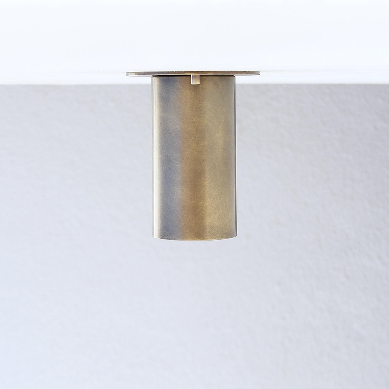 May include: A cylindrical furniture leg with a brushed brass finish. The leg features a flat circular mounting plate at the top, designed for attachment to a flat surface. The metal surface has a muted, matte texture with subtle vertical grain.