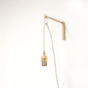 May include: A brass wall-mounted lamp arm with a hook holding a light-colored twisted fabric cord and a brass light bulb socket. The metal arm attaches to the wall with a vertical plate and extends outward to support the hanging light fixture.