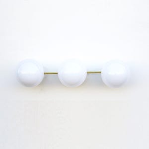 May include: A modern wall sconce featuring three white, spherical light fixtures aligned horizontally on a brass rod. The minimalist design offers a clean, contemporary aesthetic, suitable for various interior styles.
