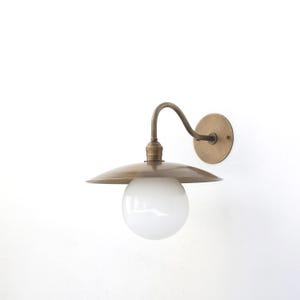 May include: A wall-mounted light fixture featuring a curved brass arm, a circular brass wall plate, a flat brass shade, and a spherical white glass globe. The metal components have a brushed finish, and the light is oriented downwards.