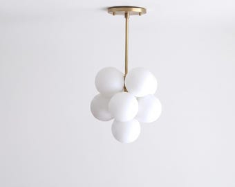 May include: A ceiling-mounted light fixture featuring a central brass-toned rod and circular canopy. The fixture holds six spherical, opaque white glass globes arranged in a cluster formation, extending downward from the brass-toned hardware.