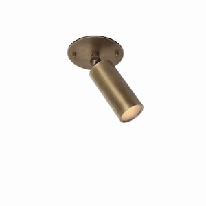 May include: A bronze-colored, cylindrical spotlight fixture with a circular base. The light is angled downward, with a warm glow emanating from the opening. The fixture is designed for ceiling mounting.