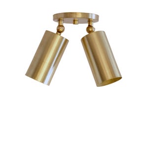 Op de afbeelding: Een plafondlamp met twee koppen, voorzien van een ronde messing montageplaat en twee cilindrische spots. Elke lichtkop is met een draaikoppeling aan de basis bevestigd, waardoor de goudkleurige lampen in verschillende standen verstelbaar zijn.