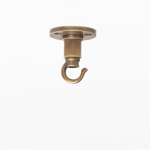 May include: A bronze-colored ceiling hook with a circular mounting plate and a curved hook. The hook is designed for hanging items from the ceiling. The metal fixture has a vintage aesthetic and is suitable for various decor styles.