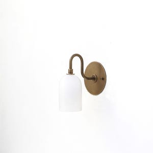 May include: A brass wall sconce with a white glass shade. The sconce has a curved arm and a round back plate.