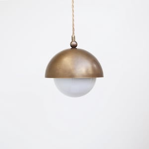 May include: A pendant light fixture with a brass-colored, semi-spherical shade and a white, globe-shaped diffuser. The light hangs from a beige cord. The fixture is a modern design, suitable for interior lighting.