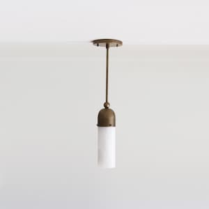 May include: A pendant light fixture with a brass finish and a white, cylindrical glass shade. The fixture is suspended from a round ceiling plate and features a slender rod. The design is minimalist and modern.