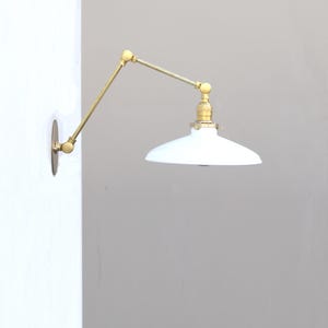 Puede incluir: Una l&aacute;mpara de pared con un brazo articulado de lat&oacute;n que cuenta con dos juntas ajustables y una placa circular. El accesorio incluye una pantalla c&oacute;nica blanca, ancha y poco profunda, unida a un casquillo de lat&oacute;n.