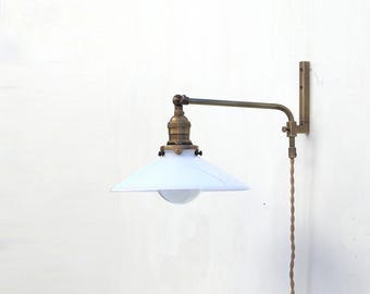 May include: A wall-mounted lamp with a white, conical shade and a brass-colored adjustable arm. The lamp has a vintage industrial design, with a visible bulb and a braided power cord. The fixture is mounted on a white wall.