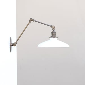 Puede incluir: Una l&aacute;mpara de pared con brazo oscilante que presenta un marco de metal de lat&oacute;n ajustable con m&uacute;ltiples articulaciones y una pantalla de metal esmaltado blanco. El accesorio se fija a la pared con una placa circular de lat&oacute;n y se extiende horizontalmente.