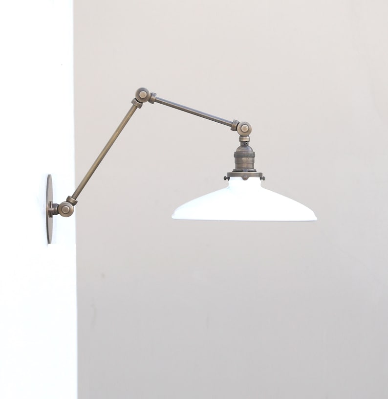 Puede incluir: Una l&aacute;mpara de pared con brazo oscilante que presenta un marco de metal de lat&oacute;n ajustable con m&uacute;ltiples articulaciones y una pantalla de metal esmaltado blanco. El accesorio se fija a la pared con una placa circular de lat&oacute;n y se extiende horizontalmente.