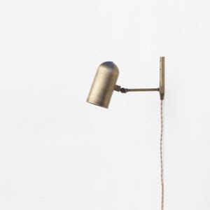 May include: A brass wall lamp with a cylindrical shade and adjustable arm. The lamp is fixed to a rectangular mounting plate and has a fabric-covered power cord. The design is minimalist and industrial, suitable for various interior styles.