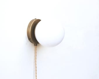 May include: A modern wall sconce featuring a round, white glass globe light. The light is mounted on a circular, antique brass-colored base. A beige, braided cord hangs down from the fixture. The sconce is designed for indoor use.