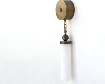May include: A wall-mounted light fixture with a cylindrical, brass-colored top and a white, cylindrical shade. The shade hangs from a chain and a brass-colored sphere. The fixture is mounted on a white wall.