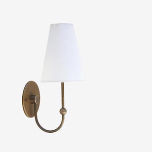 May include: A wall-mounted sconce with a white lampshade and a brass-colored metal base. The sconce features a curved arm and a round backplate. The lampshade is a classic tapered shape.