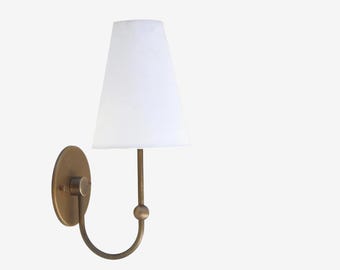 May include: A wall-mounted sconce with a white lampshade and a brass-colored metal base. The sconce features a curved arm and a round backplate. The lampshade is a classic tapered shape.