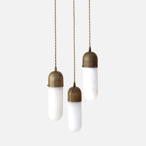 May include: Three pendant lights with a brass finish and white, capsule-shaped glass shades. The lights hang from twisted brown cords against a white background. The design is minimalist and modern.