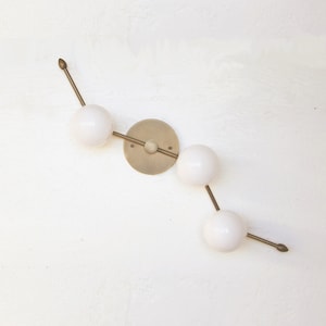 Peut inclure: Applique murale moderne avec trois globes blancs reliés par des tiges couleur laiton et une plaque de montage circulaire. L'ensemble est fixé sur un mur blanc, créant un design minimaliste et élégant.