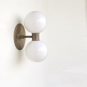 May include: A brass wall sconce with two white globe light fixtures. The sconce has a round back plate and a connecting bar between the two globes.