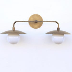 May include: A brass-toned, two-light wall sconce with a circular backplate. The fixture features a horizontal bar with two downward-facing, saucer-shaped shades. Each shade has a white, spherical light bulb.