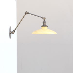 Puede incluir: Una l&aacute;mpara de pared con brazo oscilante que presenta una pantalla c&oacute;nica de color amarillo p&aacute;lido y un acabado met&aacute;lico oscuro. El accesorio incluye un brazo articulado ajustable con varias juntas, unido a una placa circular.