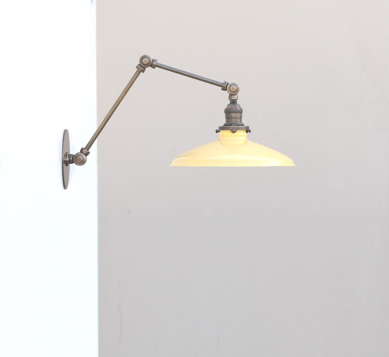 Puede incluir: Una l&aacute;mpara de pared con brazo oscilante que presenta una pantalla c&oacute;nica de color amarillo p&aacute;lido y un acabado met&aacute;lico oscuro. El accesorio incluye un brazo articulado ajustable con varias juntas, unido a una placa circular.