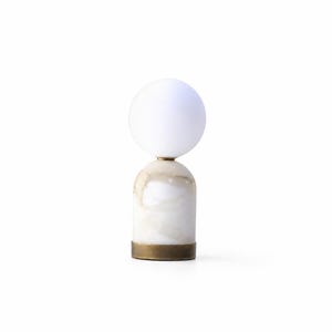 May include: A table lamp featuring a spherical white glass shade resting atop a cylindrical marble base with natural beige veining. The base is finished with a circular metallic brass-toned band at the bottom, providing a stable foundation for the light.