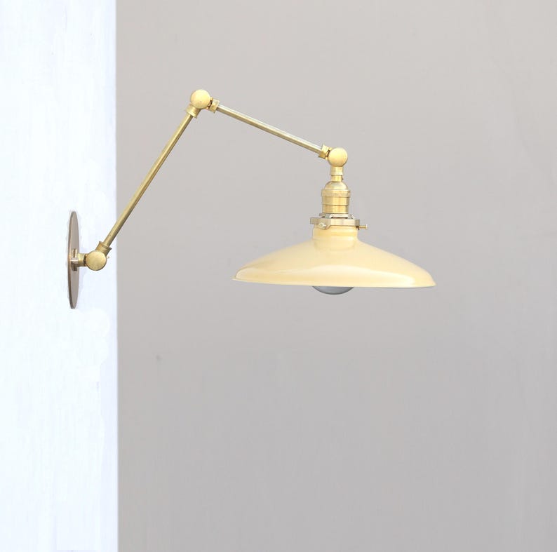 Puede incluir: Una l&aacute;mpara de pared con un brazo articulado de lat&oacute;n con m&uacute;ltiples juntas y una pantalla de metal ancha de color crema. El accesorio se fija a la pared con una placa circular de lat&oacute;n e incluye un casquillo met&aacute;lico para la bombilla.
