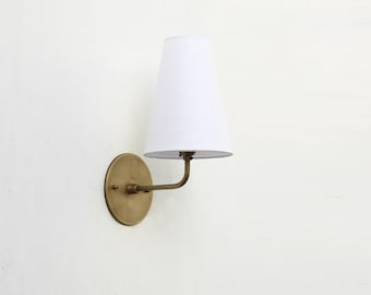 May include: A wall-mounted sconce with a white lampshade and a brass-colored metal base. The lampshade is cone-shaped, and the metal base has a circular backplate and a curved arm. The sconce is designed for indoor lighting.