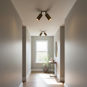 Op de afbeelding: Twee plafondlampen, elk voorzien van een centrale ronde voet met twee verstelbare cilindrische spots in een metallic bronskleurige afwerking. De lampen zijn in een gang geplaatst en werpen een warme gloed naar beneden vanaf het plafond.