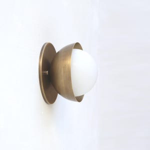 May include: A brass wall sconce featuring a white globe light. The sconce has a circular base and a half-sphere shade that surrounds the globe. The fixture is mounted on a white wall.