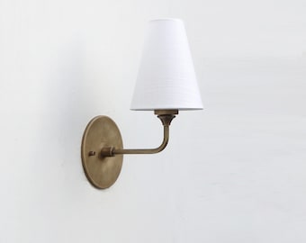 May include: A wall-mounted lamp with a white, tapered lampshade. The lamp features a brass-colored, circular base and a curved arm. The fixture is designed for indoor lighting and adds a touch of elegance to any space.