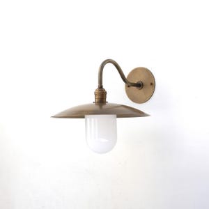 May include: Brass wall sconce with a curved arm and a white frosted glass shade. The sconce has a round, flat back plate.