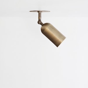 May include: A cylindrical brass spotlight fixture with a brushed finish, featuring a circular ceiling mount and an adjustable swivel joint that allows the light head to be angled downward for directional illumination in a room.