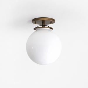 May include: A flush mount ceiling light fitting featuring a large, spherical white glass globe shade attached to a circular, antique brass-finished metal base. The hardware includes a decorative metal ring that secures the glass globe to the mount.