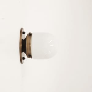 May include: A brass wall sconce with a white glass shade. The sconce has a round base and a decorative metal band around the shade.