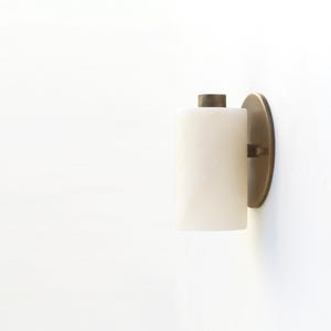 May include: A modern wall sconce with a cylindrical, off-white shade and a brass-colored backplate. The sconce is mounted on a white wall, providing a clean and minimalist aesthetic. The fixture is designed for indoor lighting.