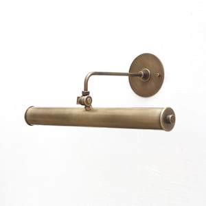 May include: A brass picture light with a long, cylindrical shade and a round wall mount. The light has a swivel arm that allows it to be adjusted to direct the light.