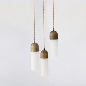 May include: Three pendant lights with cylindrical white glass shades and antique brass-colored metal caps. The lights hang from twisted tan cords against a white background. The design is minimalist and modern.