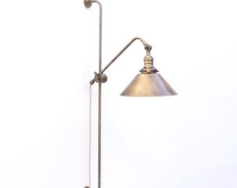 May include: A wall-mounted lamp with a brass finish, featuring a long vertical rod, an adjustable swing arm, and a conical shade. The fixture includes a braided fabric cord that hangs down from the central joint, providing a functional lighting solution.