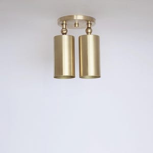 Op de afbeelding: Een plafondlamp met twee lichtpunten, voorzien van een ronde messing montageplaat en twee cilindervormige messing kappen. Elke kap is aan de basis bevestigd met een klein kogelgewricht, waardoor de richting van het licht kan worden aangepast.