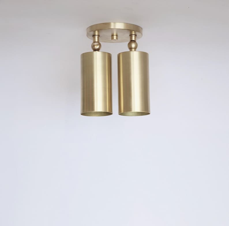 Op de afbeelding: Een plafondlamp met twee lichtpunten, voorzien van een ronde messing montageplaat en twee cilindervormige messing kappen. Elke kap is aan de basis bevestigd met een klein kogelgewricht, waardoor de richting van het licht kan worden aangepast.