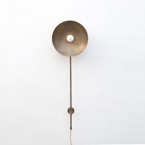 May include: A brass wall sconce with a circular shade and a white bulb. The sconce has a long, slender arm and a small circular base. The design is minimalist and modern, suitable for interior lighting.