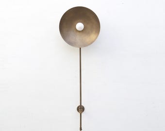 May include: A brass wall sconce with a circular shade and a white bulb. The sconce has a long, slender arm and a small circular base. The design is minimalist and modern, suitable for interior lighting.