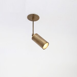 May include: A modern, brass-toned ceiling spotlight fixture. The cylindrical light head is angled, emitting a warm glow. The fixture includes a round ceiling mount and a slender connecting rod. This minimalist design is suitable for various interior styles.