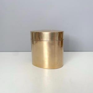 May include: A gold-colored, cylindrical container with a lid. The container has a smooth, reflective surface and sits on a white surface. The lid fits snugly on top, creating a seamless look. The container is likely used for storage.