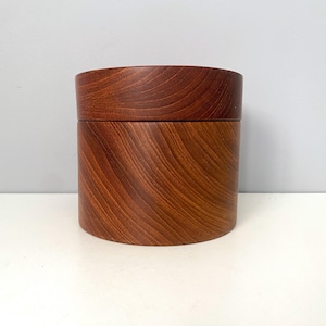 May include: A cylindrical wooden box with a lid. The box and lid are made of a reddish-brown wood, with visible grain patterns. The box is sitting on a white surface against a light gray background.