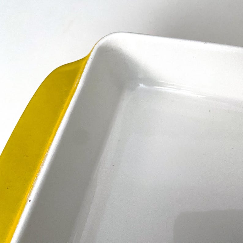 Op de afbeelding: Close-up van een rechthoekige witte ovenschaal met een felgele rand. De schaal heeft een glanzende afwerking en een eenvoudig, modern ontwerp. De hoek van de schaal is zichtbaar tegen een witte achtergrond.